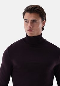 Junger Mann mit dunklen Haaren, der einen schwarzen Rollkragenpullover trägt und leicht seitlich blickt vor einem schlichten hellen Hintergrund.