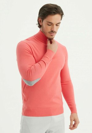 Pull en col roulé en tricot doux, avec des patchs de coude gris et des manches ajustées, associé à un pantalon de couleur claire.