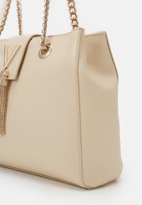 Beige strukturierte Handtasche mit einer Kettenriemen, goldfarbenen Metallteilen, einem Quastenakzent und einer strukturierten Form mit einer Klappenverschluss.