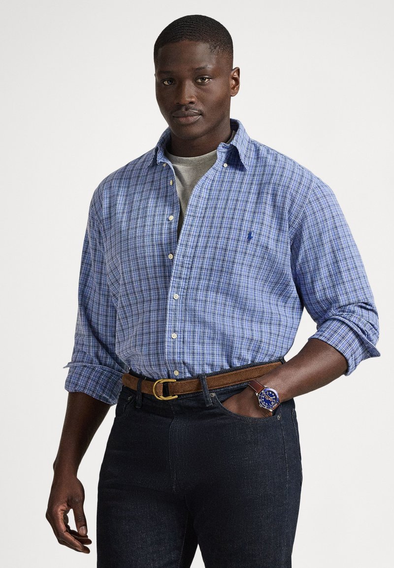 Modro-bela srajca s kockami, z zavihani rokavi, kombinirana s temnimi kavbojkami, rjavim pasom in modro uro z usnjenim paščkom.