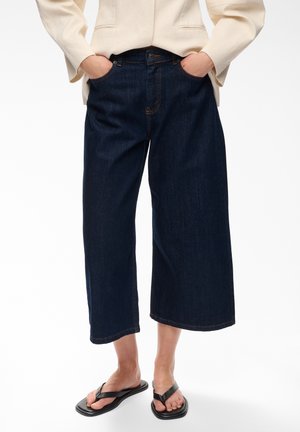 Personne portant un jean court large de couleur foncée, un blazer crème avec les mains dans les poches, et des sandales flip-flop noires sur fond blanc.