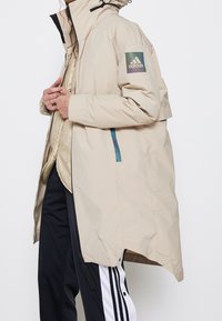 Beżowa wodoodporna parka z wysokim kołnierzem, regulowanym kapturem, bocznymi kieszeniami na zamek oraz naszywką z logo Adidasa na lewym rękawie.