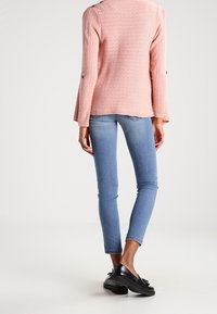 Rosa strukturiertes Sweatshirt mit ausgestellten Ärmeln, kombiniert mit hellblauen figurbetonten Jeans und schwarzen Schuhen, die ein lässiges Outfit betonen.