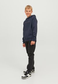 Felpa da marina con tasca frontale e testo, abbinata a jeans neri e scarpe da ginnastica bianco e nere. Tessuto morbido e vestibilità casual.