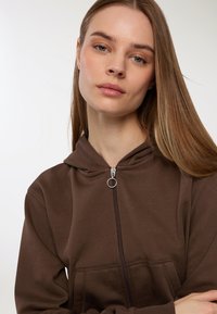 Giovane donna con lunghi capelli castano chiaro e lisci, che indossa una felpa con zip marrone, guarda con calma la camera su uno sfondo neutro.