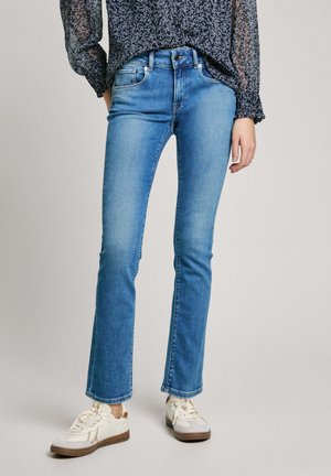 Femme portant un jean droit bleu clair, un chemisier fleuri transparent à manches longues et des baskets blanches avec des semelles brunes.