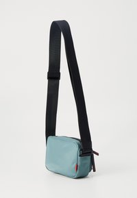 Geantă mică și rectangulară pentru umăr, din țesătură de culoare teal deschis, având o curea neagră reglabilă și un accent de marcă roșu pe lateral. Închidere cu fermoar.