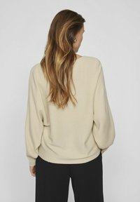 Beżowy sweter prążkowany o luźnym kroju, z szerokimi rękawami i zaokrąglonym dekoltem na plecach, w zestawieniu z czarnymi spodniami o luźnym kroju.