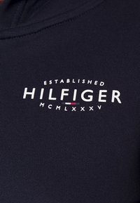 Tissu bleu marine avec texte blanc "HILFIGER", petit logo rouge, blanc et bleu en dessous, et "ESTABLISHED MCMLXXXV" imprimé au-dessus et en dessous.