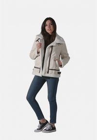 Veste en peau lainée blanche avec grand col, accents marron et poches zippées. Associée à un jean bleu et des baskets noires.