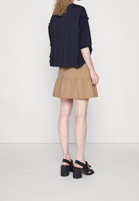 Donna che indossa un top blu navy con dettagli a volant, una gonna flare beige e sandali neri con tacco a blocco, in piedi contro uno sfondo bianco.