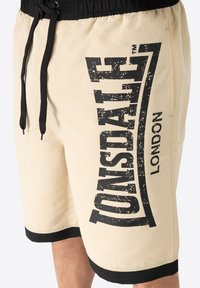 Beige shorts with a black elastic waistband, featuring a large distressed black logo print on the side and black hem accents.