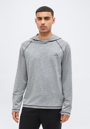 Άνδρας που φοράει ανοιχτό γκρι αθλητική μπλούζα με μακριά μανίκια και διακριτικό λογότυπο Adidas, στέκεται μπροστά σε απλό λευκό φόντο.