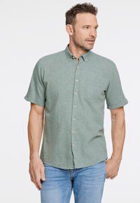 Camicia button-up a maniche corte verde chiaro con tessuto testurizzato, tasca sul petto singola e bottoni marroni, indossata con jeans blu.