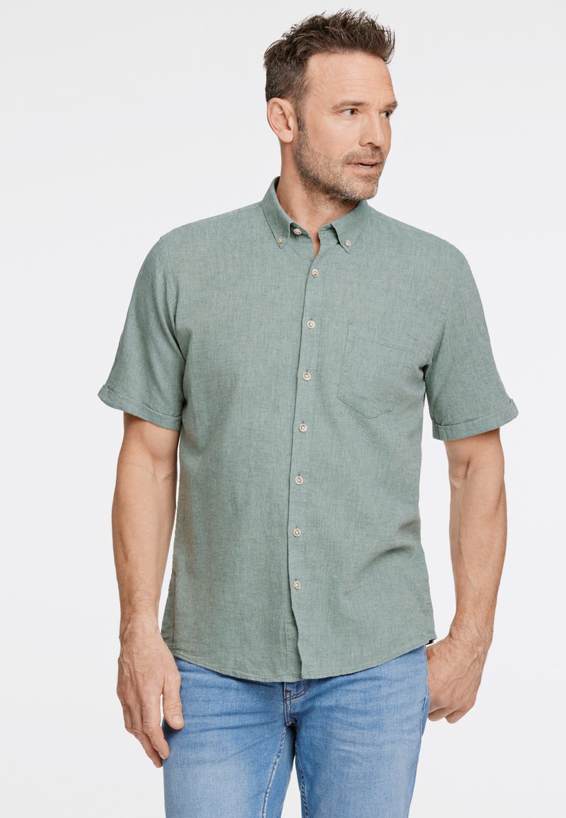 Camicia button-up a maniche corte verde chiaro con tessuto testurizzato, tasca sul petto singola e bottoni marroni, indossata con jeans blu.