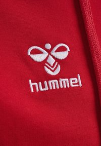 Rode stof met een gladde textuur, met een wit geborduurd logo en de tekst "hummel" onder een gestileerd ontwerp, prominent weergegeven.