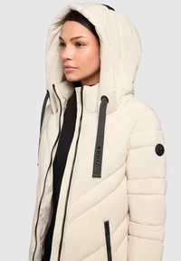 Giacca puffer crema con cappuccio, caratterizzata da cuciture diagonali, tasche con zip nere e un'etichetta con logo sul laccio del cappuccio.