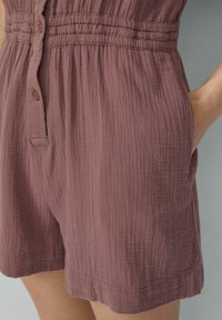 Mauve Romper mit Puffärmeln aus strukturiertem Stoff, ausgestattet mit einer Knopfleiste und elastischem Bund, dazu seitliche Taschen für zusätzlichen Komfort.