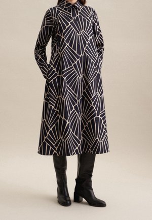 Robe chemise bleu marine à motifs géométriques crème, manches longues, coupe décontractée, associée à des bottes noires montantes jusqu'aux genoux.