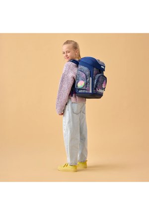 Mädchen trägt einen lila Pullover und helle Jeans, hat einen großen blauen Rucksack mit Blumen-Patches und steht vor einem beigen Hintergrund.