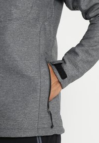 Veste en polaire grise avec une poche latérale zippée et fermeture à velcro aux poignets. Texture douce avec un design lisse et moderne.