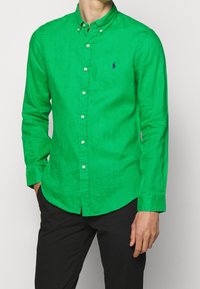 Grön långärmad skjorta med knappar i en texturerad tyg, med krage och en liten logo på vänster bröst. Bärs med mörka byxor.