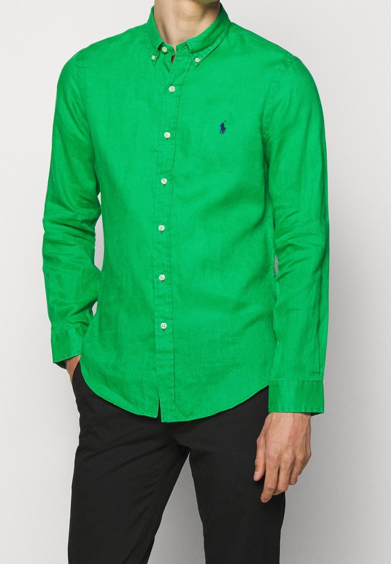 Grön långärmad skjorta med knappar i en texturerad tyg, med krage och en liten logo på vänster bröst. Bärs med mörka byxor.