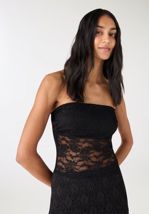 Kobieta o długich ciemnych włosach, ubrana w czarną koronkową sukienkę bez ramiączek z przezroczystą koronkową częścią w talii, pozująca na jednolitym jasnym tle.