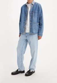 Man med ljusblå denimjeans, svarta läderskor, vit t-shirt, randig skjorta med knappar och en blå denimjacka med lapplösa fickor.