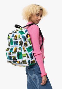 Vit tygryggsäck med ett collage av färgglada rese-teman tryck. Har svarta remmar och en framficka med dragkedja. Standard ryggsäcksform.