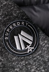 Tessuto a maglia grigio con un logo ricamato circolare che mostra "SUPERDRY" e disegni geometrici in bianco e nero. Dettagli della superficie testurizzata visibili.