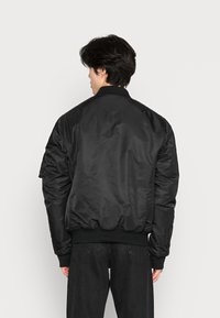 Giacca bomber nera con una texture liscia e lucida, polsini e orlo a coste, e una vestibilità rilassata. Presenta una tasca utility sulla manica sinistra.