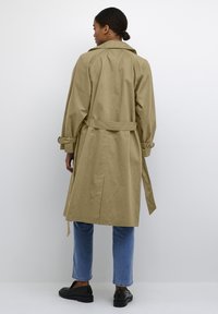 Khakifarbenes Trenchcoat mit tailliertem Gürtel, langen Ärmeln und klassischem Kragen. Kombiniert mit blauen Jeans und schwarzen Slippern.