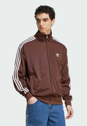Brūns zip-up sporta jaka ar augstu apkakli, baltām trīs svītrām uz piedurknēm un izšūtu logotipu uz krūtīm.