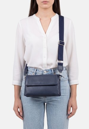 Donna che indossa una camicetta bianca e jeans azzurri con una borsa a tracolla in pelle blu scuro con tracolla regolabile.