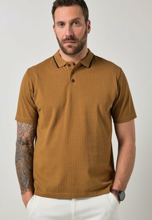 Uomo con barba che indossa una polo marrone a motivo, pantaloni bianchi, orologio da polso e un tatuaggio a manica dettagliato sul braccio sinistro, in piedi con le mani nelle tasche.