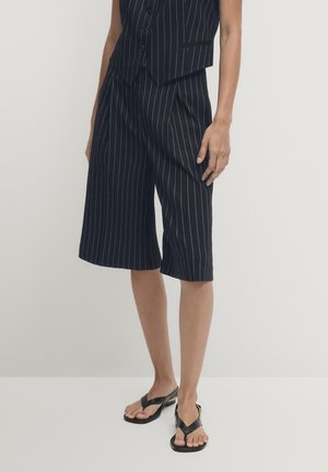 Persoon draagt zwarte, brede pinstripe korte broek en bijpassend vest, gecombineerd met zwarte teenslippers, staand tegen een effen achtergrond.