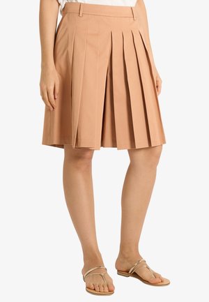 Donna che indossa una gonna plissettata beige al ginocchio con passanti per cintura abbinata a sandali infradito beige su sfondo bianco.