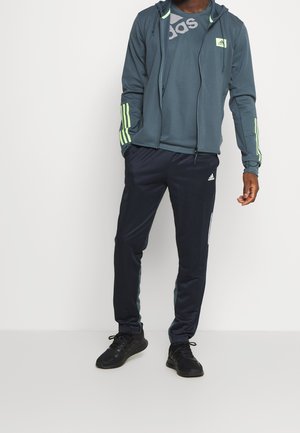 Trainingspakset met een donkergroene ritsjack met gele accenten en een grafisch T-shirt, gecombineerd met marineblauwe broek en zwarte sportschoenen.