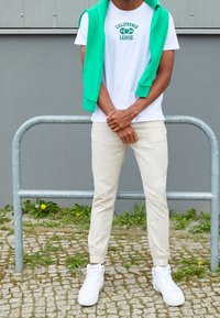 Vit bomullst-shirt med grön text, kombinerad med beiga joggingbyxor och vita höga sneakers. En grön hoodie är slängd över axlarna.