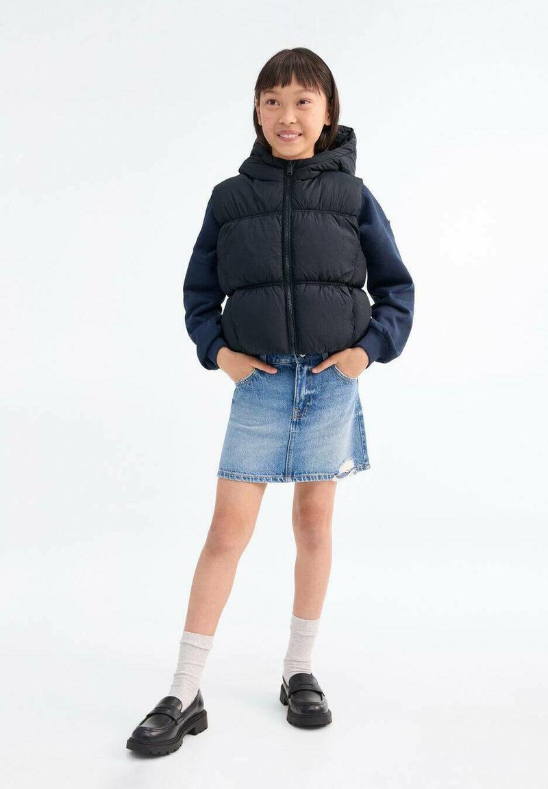 Črn puhovka z kapuco čez temno dolgo majico, v kombinaciji s svetlo modrim jeans krilom in črnimi mokasini; vidne svetlo sive nogavice.