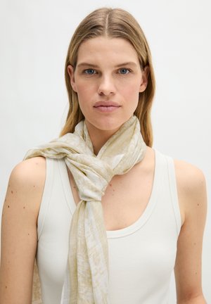 Vrouw met steil blond haar die een licht mouwloos topje draagt en een losjes geknoopte beige sjaal met patroon rond haar nek.