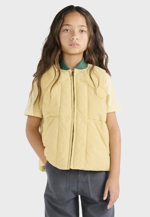 Gilet imbottito giallo chiaro con maniche corte e zip frontale. Presenta un colletto verde e due tasche frontali. Indossato sopra una camicia color crema.