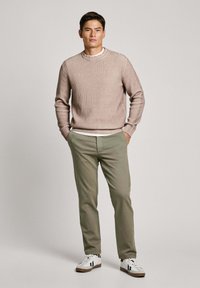 Beige ribbed sweater dengan leher bulat, dipasangkan dengan celana hijau zaitun dan sepatu sneaker putih dengan aksen hitam. Model berdiri dengan percaya diri.