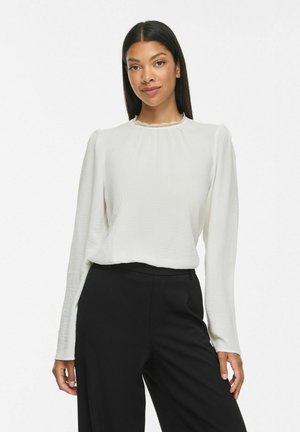 Femme aux longs cheveux noirs portant un chemisier blanc à manches longues avec un col en dentelle et un pantalon noir, debout devant un fond blanc uni.