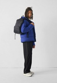 Chaqueta acolchada azul con cremallera, mochila negra, pantalones negros con costuras blancas y zapatillas beige. Fondo liso.