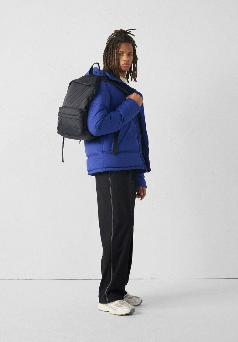Chaqueta acolchada azul con cremallera, mochila negra, pantalones negros con costuras blancas y zapatillas beige. Fondo liso.