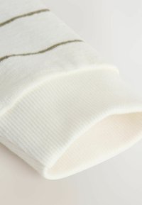Biela pletená manžeta svetra s ryhovanou textúrou a horizontálnymi olivovými pruhmi. Mäkká tkanina, hladký povrch, zúžený dizajn.