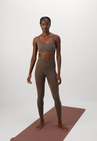 Mulher em pé descalça sobre um tapete de yoga castanho, vestindo um top desportivo castanho a condizer e leggings de cintura alta, contra um fundo branco simples.
