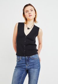 Svart ermeløs cardigan med V-hals og seks knapper, kombinert med blå denimjeans, satt mot en enkel hvit bakgrunn.